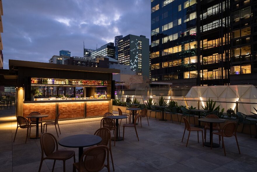 The rooftop bar at Bossley at night, with an illuminated bar service area, tables and chairs arranged on a patio, and views of city  buildings in the background.