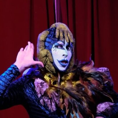 Drag performer wearing white and purple makeup and catsuit.