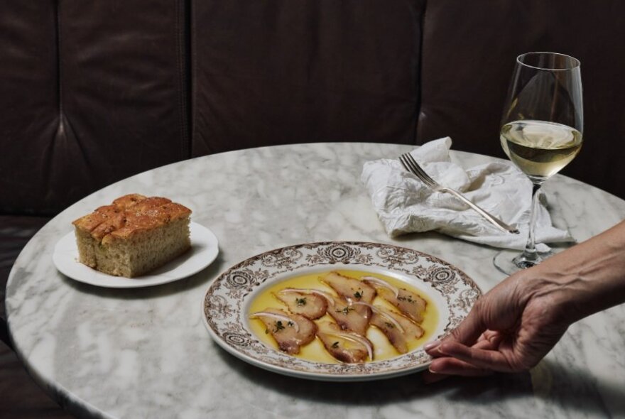 Waiter's hand placing a plate of cod on a marble table with a slice of cake and glass of white wine.