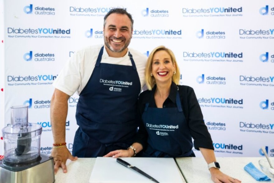 Two chefs standing wearing aprons, standing behind a bench with cooking utensils and chopping board.