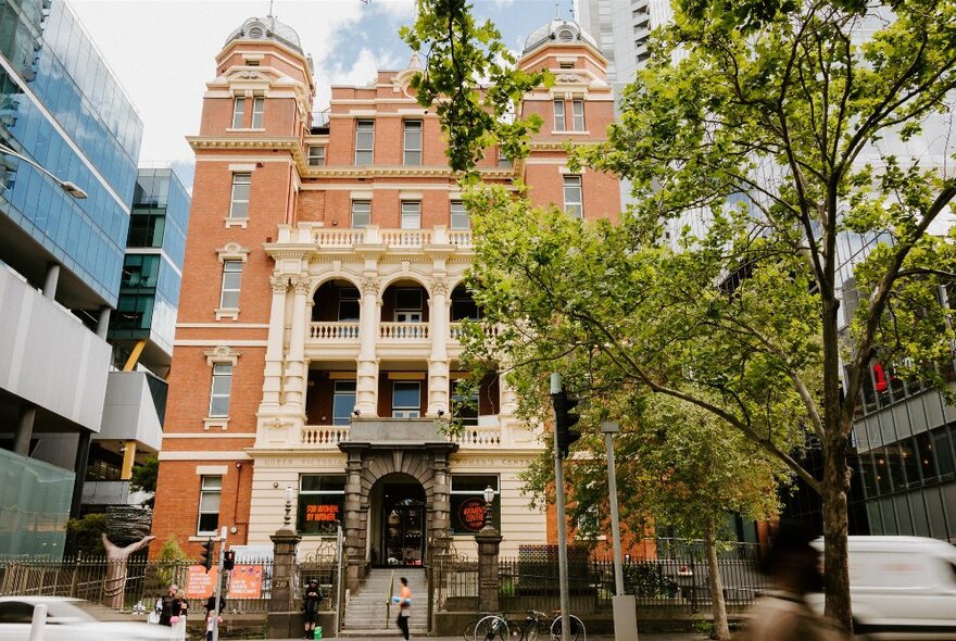 Red and cream brick multi-storied exterior of the Queen Victoria Women's Centre with modern city buildings on either side, a leafy tree and pedestrians in the foreground at street level.