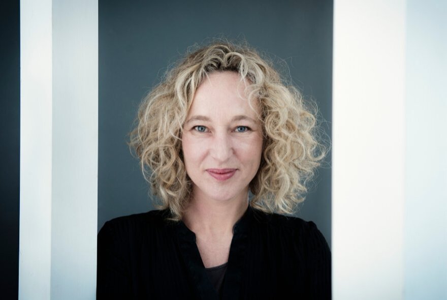 Sally Warhaft, a woman with wavy blonde hair wearing a dark top, posed against a dark grey background.