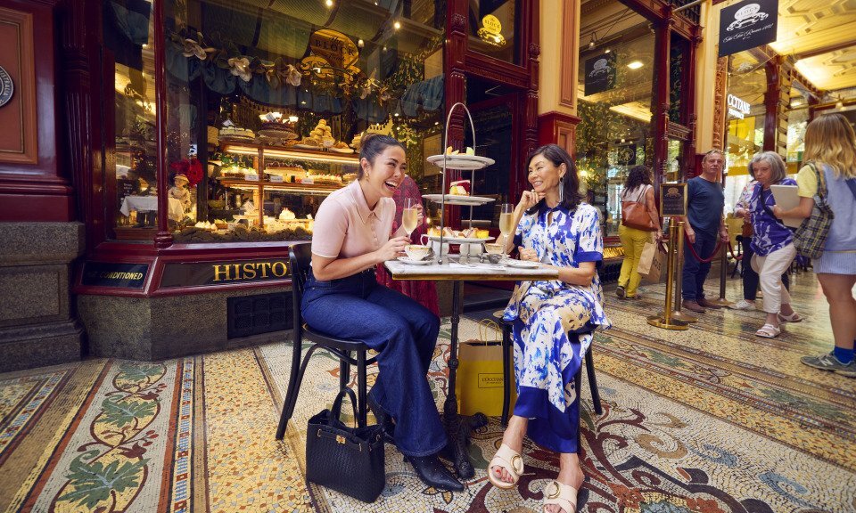 Two women laughing while they sit inside an arcade having high tea.