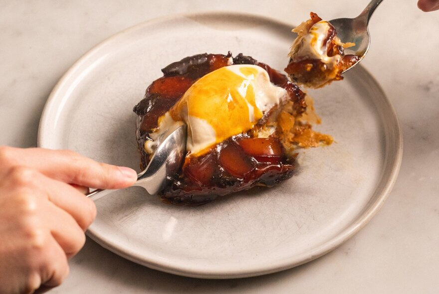 A person using a spoon to scoop into an individual tarte tatin topped with melting vanilla ice cream on a light-coloured plate.