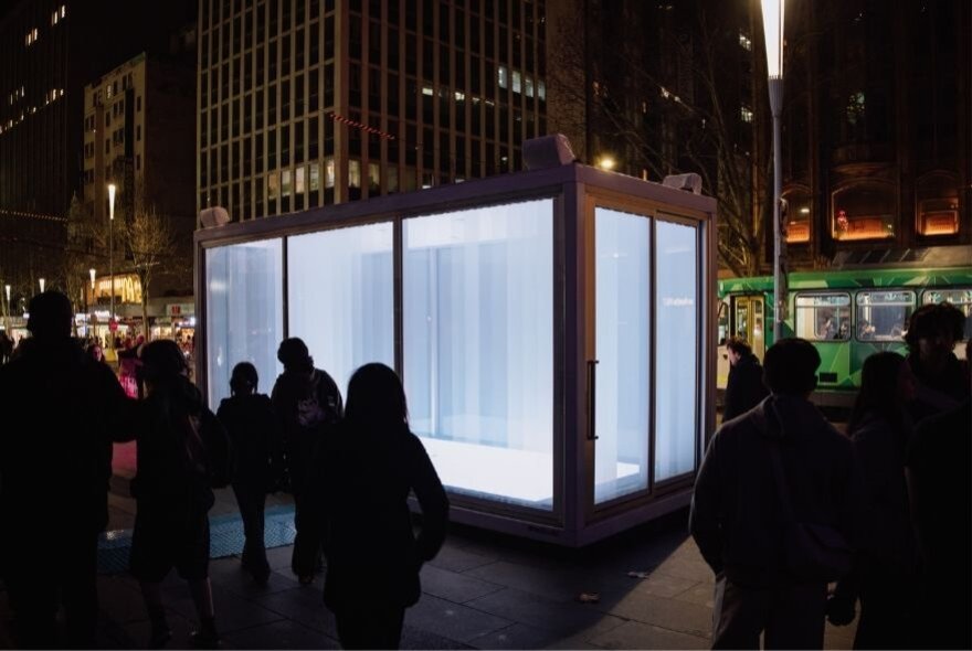 A large clear box in a Melbourne street, lit up from the inside at night.