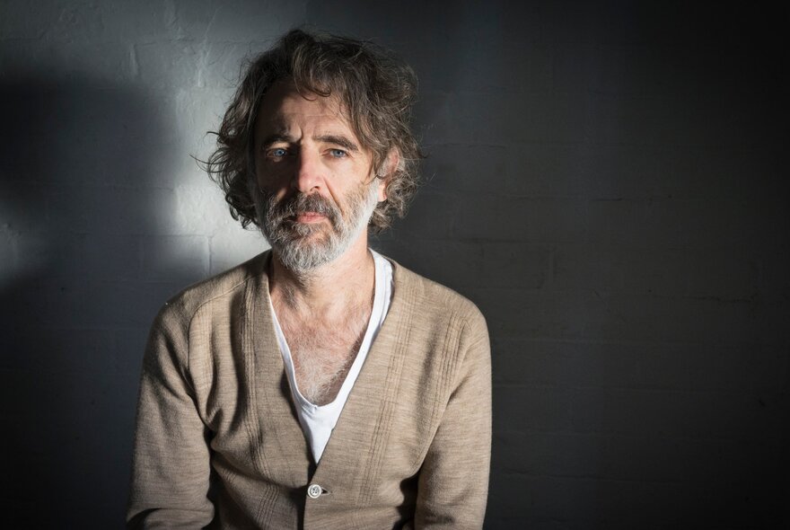 Author Dean Manning, a middle-aged man with scruffy hair and a beard, wearing a tan cardigan and seated in front of a dark wall.