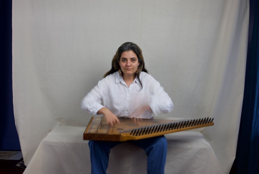 A seated person playing a qanun, a traditional Middle Eastern stringed instrument, against a white background.