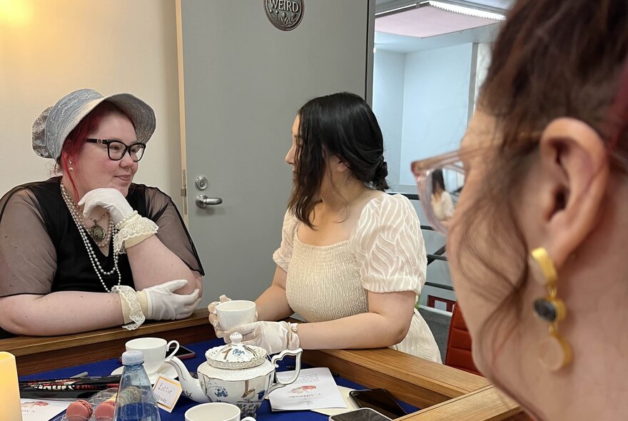 Three women in Regency-era themed costumes enjoy tea at a table.