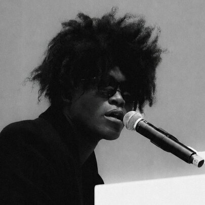 British musician Benjamin Clementine wearing dark glasses, he has an Afro hairdo and performs and sings at a piano.