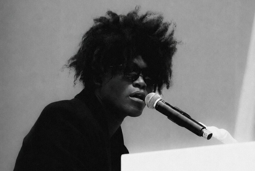 British musician Benjamin Clementine wearing dark glasses, he has an Afro hairdo and performs and sings at a piano.
