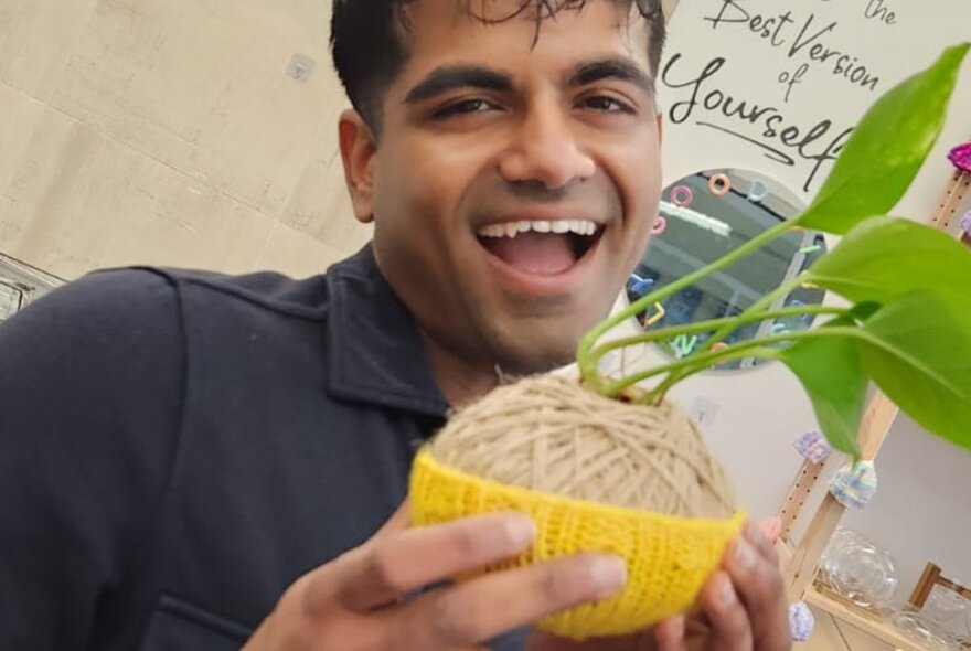 A man smiling with an open mouth, holding up a kokedama moss ball he has made at a workshop.