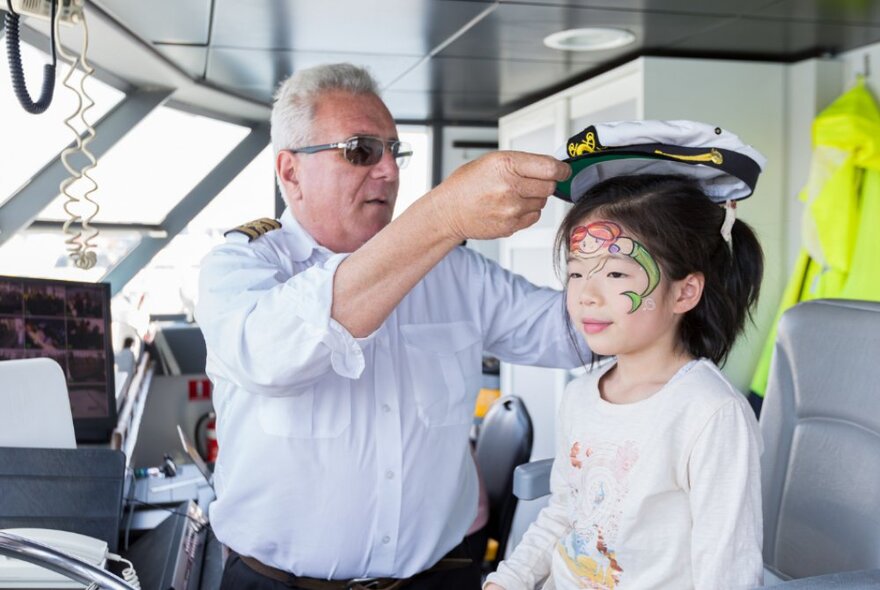 A boat captain placing a captain's cap on the head of a child who sits on the cockpit of a boat.