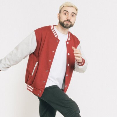 Comedian Simon Taylor, a light-skinned man with short blonde hair and a dark beard, wearing a red and white varsity jacket over a white t-shirt and black pants, captured in a mid-stride pose.