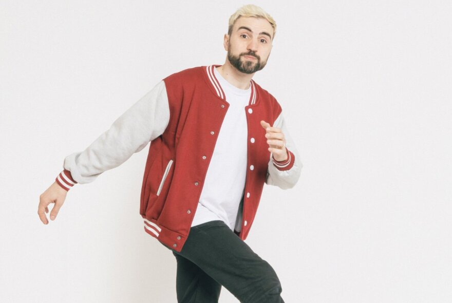 Comedian Simon Taylor, a light-skinned man with short blonde hair and a dark beard, wearing a red and white varsity jacket over a white t-shirt and black pants, captured in a mid-stride pose.