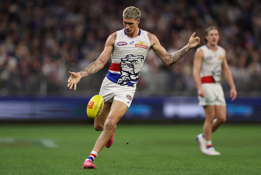 A Western Bulldogs AFL player kicking a yellow football during a game.