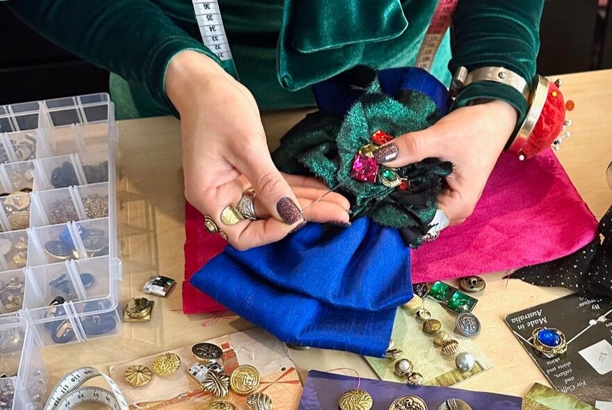Looking down at hands crafting a large fabric bow, buttons and beads scattered around on the table. 