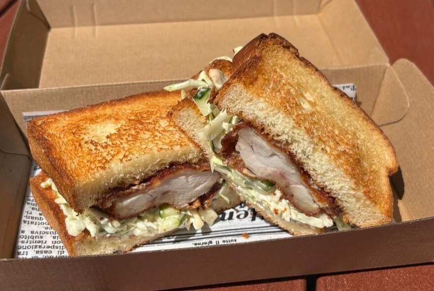 A toasted sandwich with chicken schnitzel and coleslaw fillings, inside a brown cardboard box.