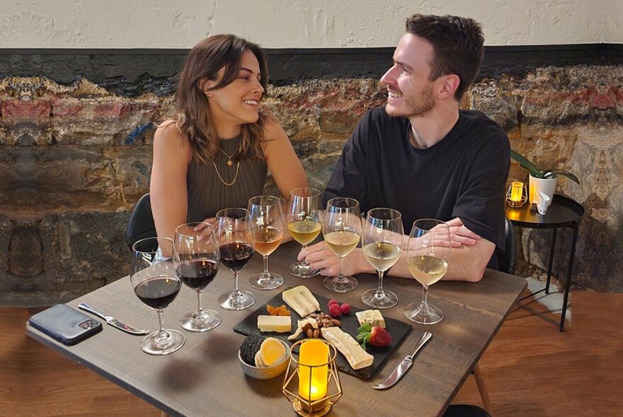 Two people seated at a dining table with a flight of wine glasses and a cheese platter in front of them.