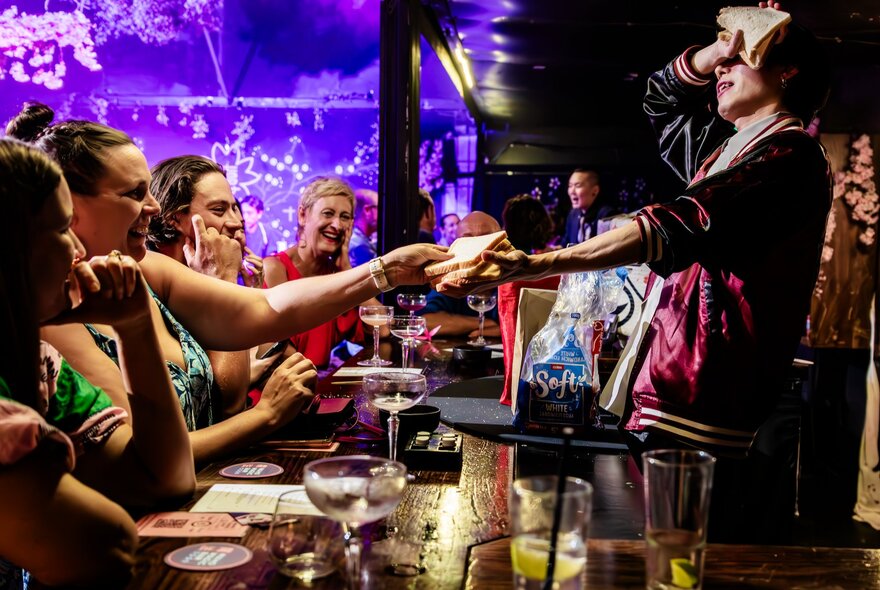 Performer with hand oustretched and eyes covered, seated people interacting over table.