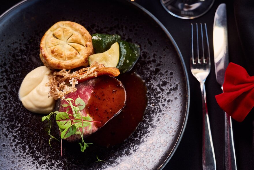 A fine-dining course featuring a piece of meat with a pastry component, purée, and a rich jus on a dark plate.