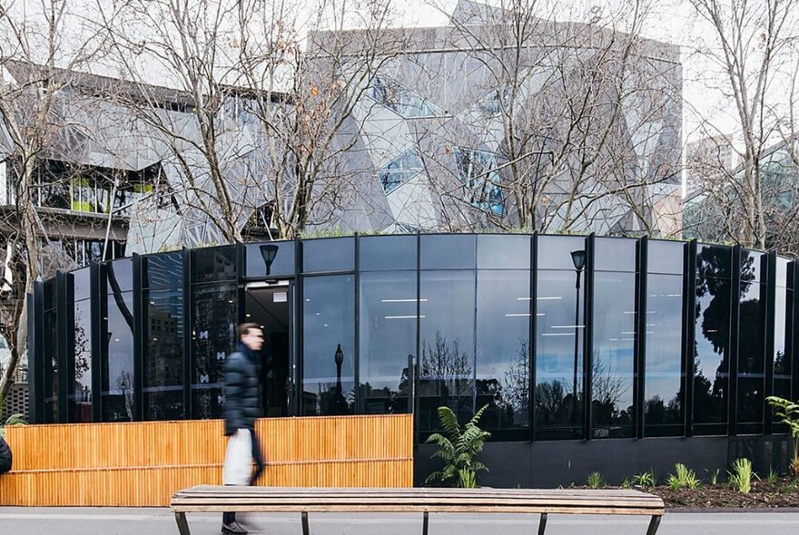 Exterior of a glass surround building with bare winter trees and a man walking by in a black puffer jacket. 