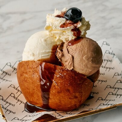 A scoop of gelato in a bun with whipped cream, a maraschino cherry and fudge on top.