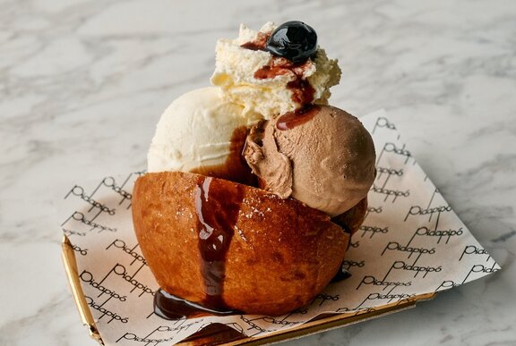A scoop of gelato in a bun with whipped cream, a maraschino cherry and fudge on top.