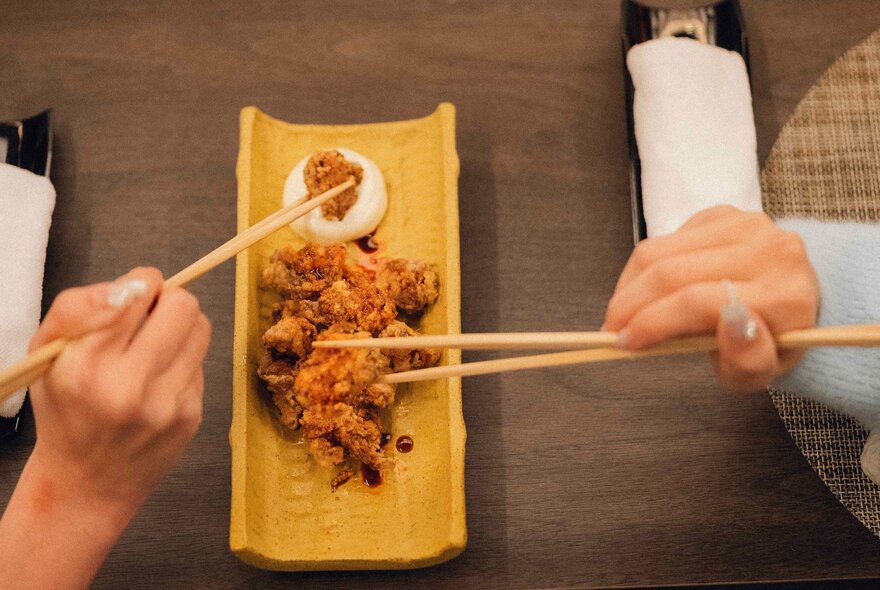 Two people's hands using chopsticks to pick up pieces of karaage, Japanese fried chicken, off a yellow platter. 