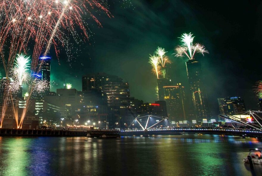 Fireworks exploding in the night sky over Melbourne city buildings, with the Yarra River in the foreground.