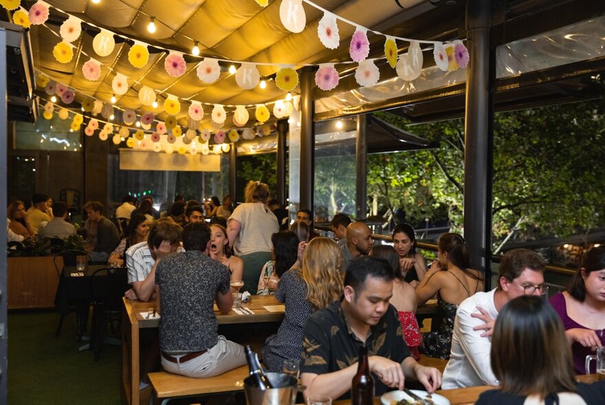 People gathered at outdoor tables under an awning lit with party lights and bunting. 