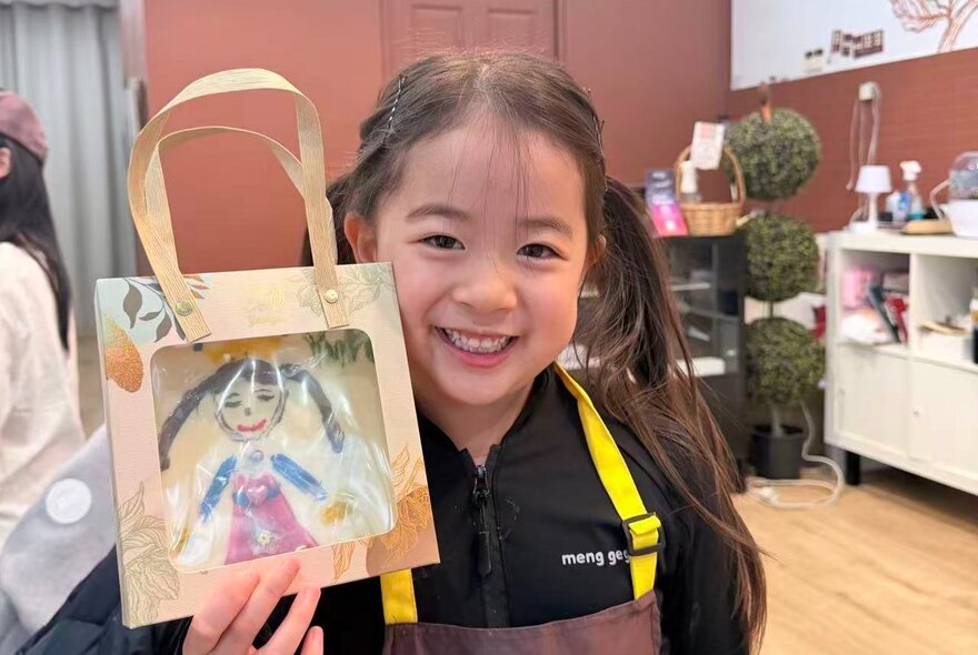 A smiling child holding up a hand-painted chocolate in a gift bag, standing inside a chocolate shop.