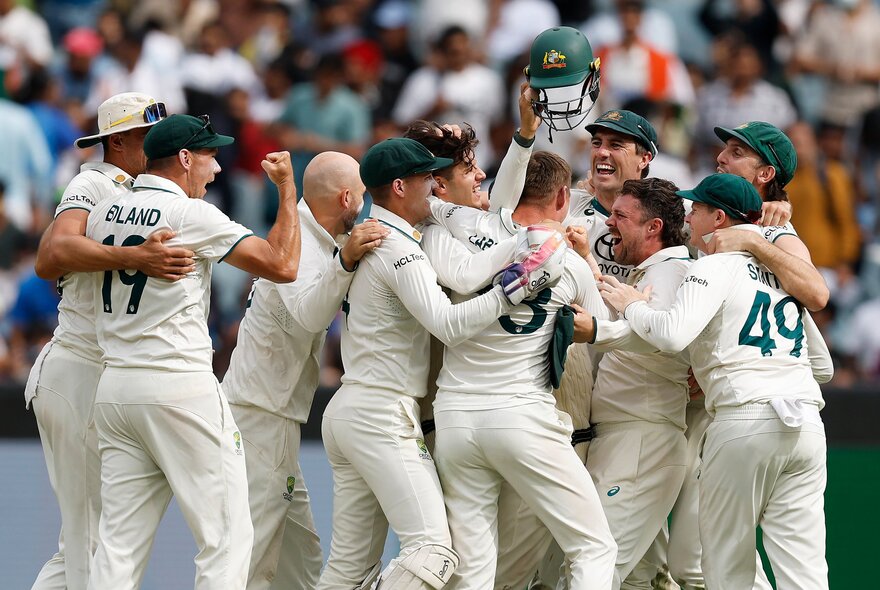 Australian cricket players, wearing their white Test uniforms, cheering and celebrating in a group huddle on the field, with out of focus sports fans in the stadium behind them.