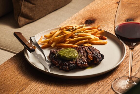 A white plate with steak and chips on a wooden table with glass of wine