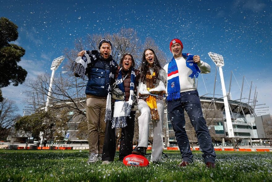 Four AFL footy fans wearing team scarves, standing in front of the MCG, with a sprinkle of snow falling around them.