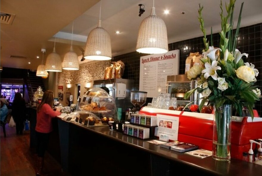 Person standing at cafe counter with flower arrangement and display cabinet of muffins and pastries.