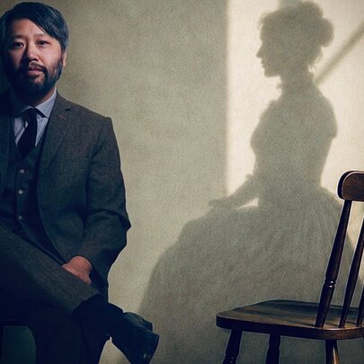 A man in a three-piece suit sitting on a stool holding a lit candle, with the shadow silhouette of a woman wearing a Victorian-style gown sitting in an empty wooden chair positioned next to him on the wall.