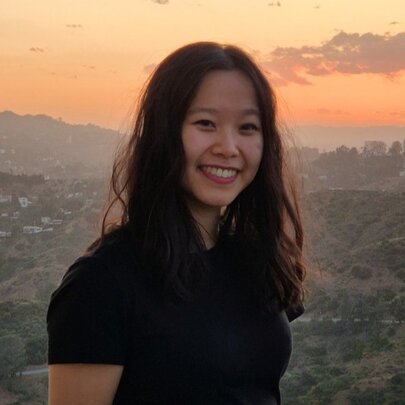 Young woman with long dark hair standing in front of a hilly background with buildings and sunset.
