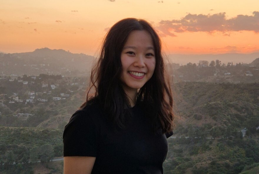 Young woman with long dark hair standing in front of a hilly background with buildings and sunset.