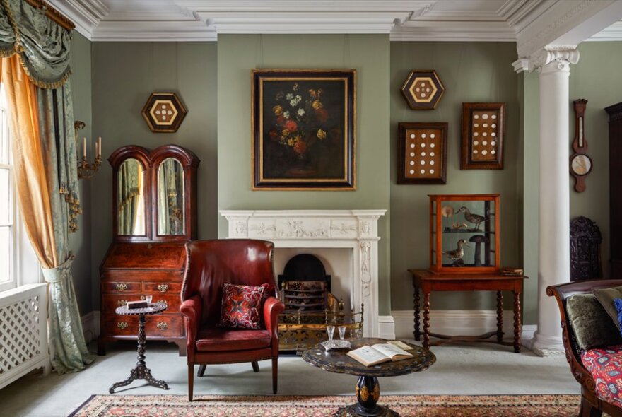 A room at The Johnson Collection Georgian mansion with a fireplace, a leather armchair, a mirrored writing bureau, rugs, objects d'art and paintings on the wall. 