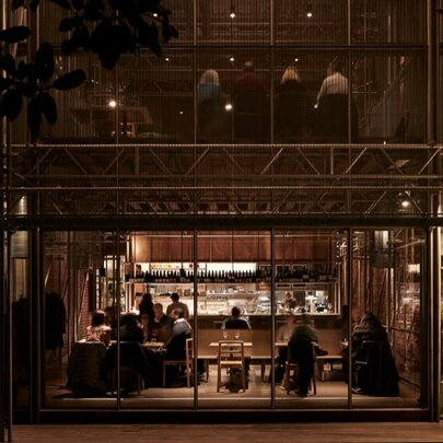The exterior glass facade of an industrial-style restaurant, with patrons seated alongside a dimly lit bar inside.