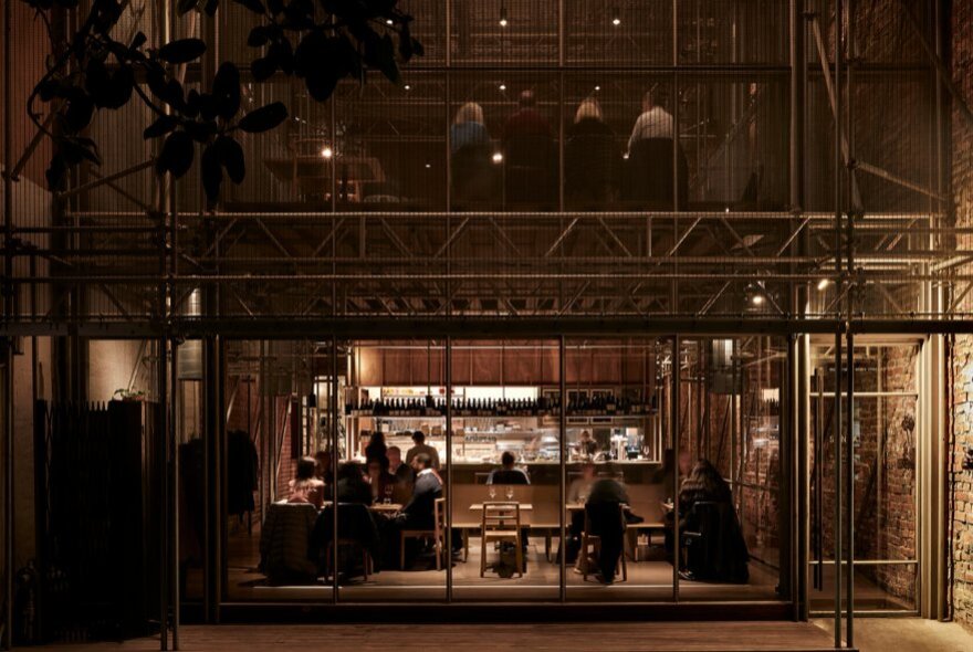 The exterior glass facade of an industrial-style restaurant, with patrons seated alongside a dimly lit bar inside.