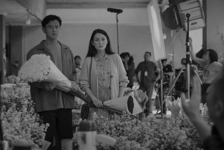 Black and white film still, a couple holding bouquets of flowers at a flower stall, camera crew in the background.