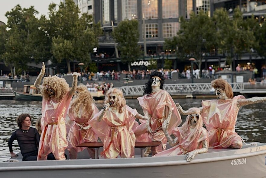 A music group wearing shining pink robes and glittering gold bodysuits performing on a small boat in the Yarra River.