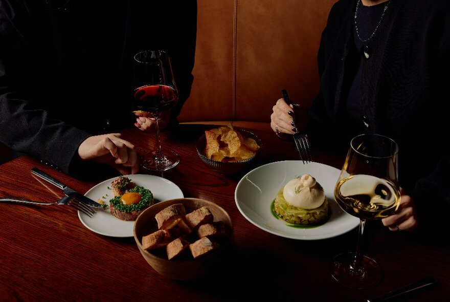 Two people dining at a booth table, with two dishes of food in front of them, and glasses of wine.