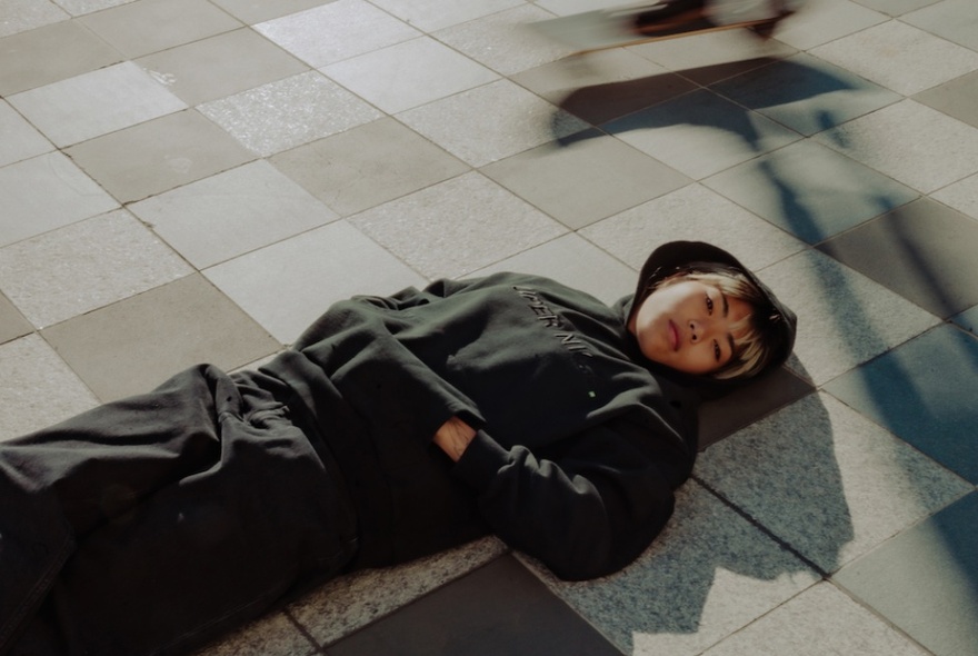 A woman wearing a dark green hooded jumper and tracksuit pants, lying down on a tiled floor with hands in the front pocket of her jumper.