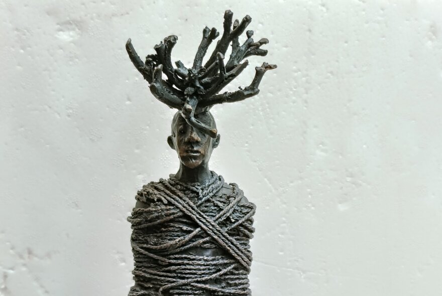 Bronze sculpture of a human figure with its torso tightly wrapped in thick ropes and a dense, antler-like crown of branches or roots  sprouting from the figure's head.