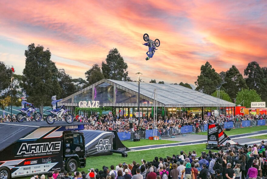 Crowds of people at the Melbourne Royal Show watching a freestyle motorbike rider performing mid-air stunts on their bike, set against an orange coloured early evening sky. 