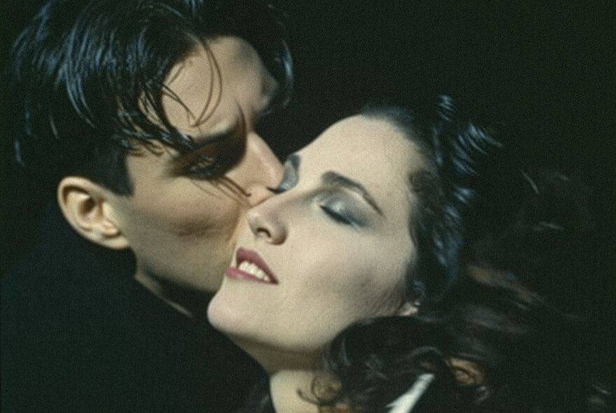 Woman with eyes closed and head back, her cheek being kissed by a man with dark eyebrows and hair.