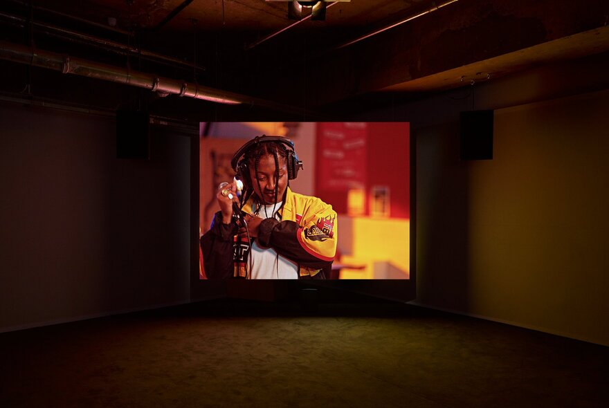 A darkened room where a projection of a person recording vocals is playing. 