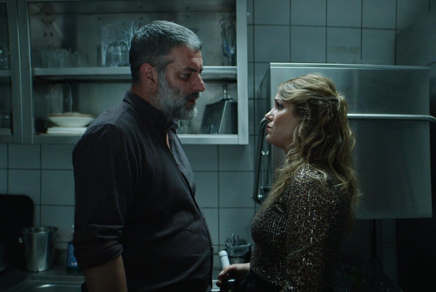 A still from a movie showing two people in side profile facing each other in a kitchen setting.